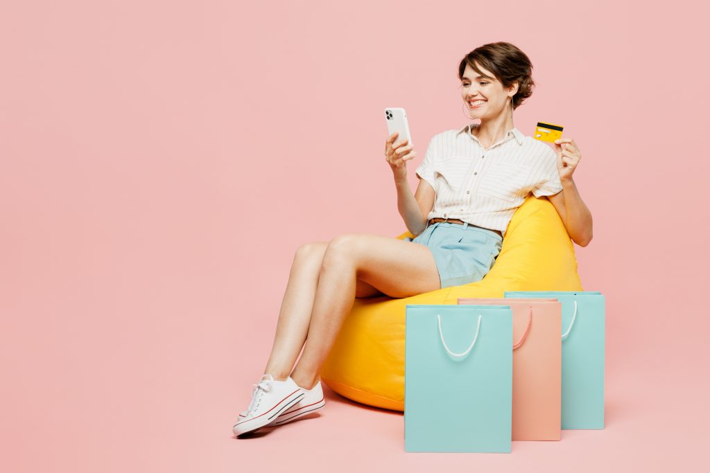 Full body happy young woman wear casual clothes hold bags sit in bag chair use mobile cell phone credit bank card shopping online isolated on plain pink background. Black Friday sale buy day concept.